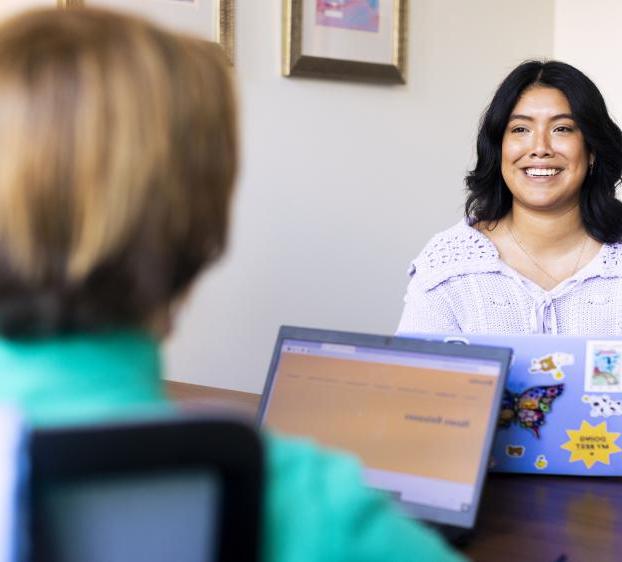 A student sits at the desk of a faculty member and smiles as they chat in the professor's office during office hours.